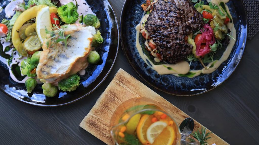 Top view of gourmet fish and steak dishes with fresh citrus and herbs on a stylish table setting.